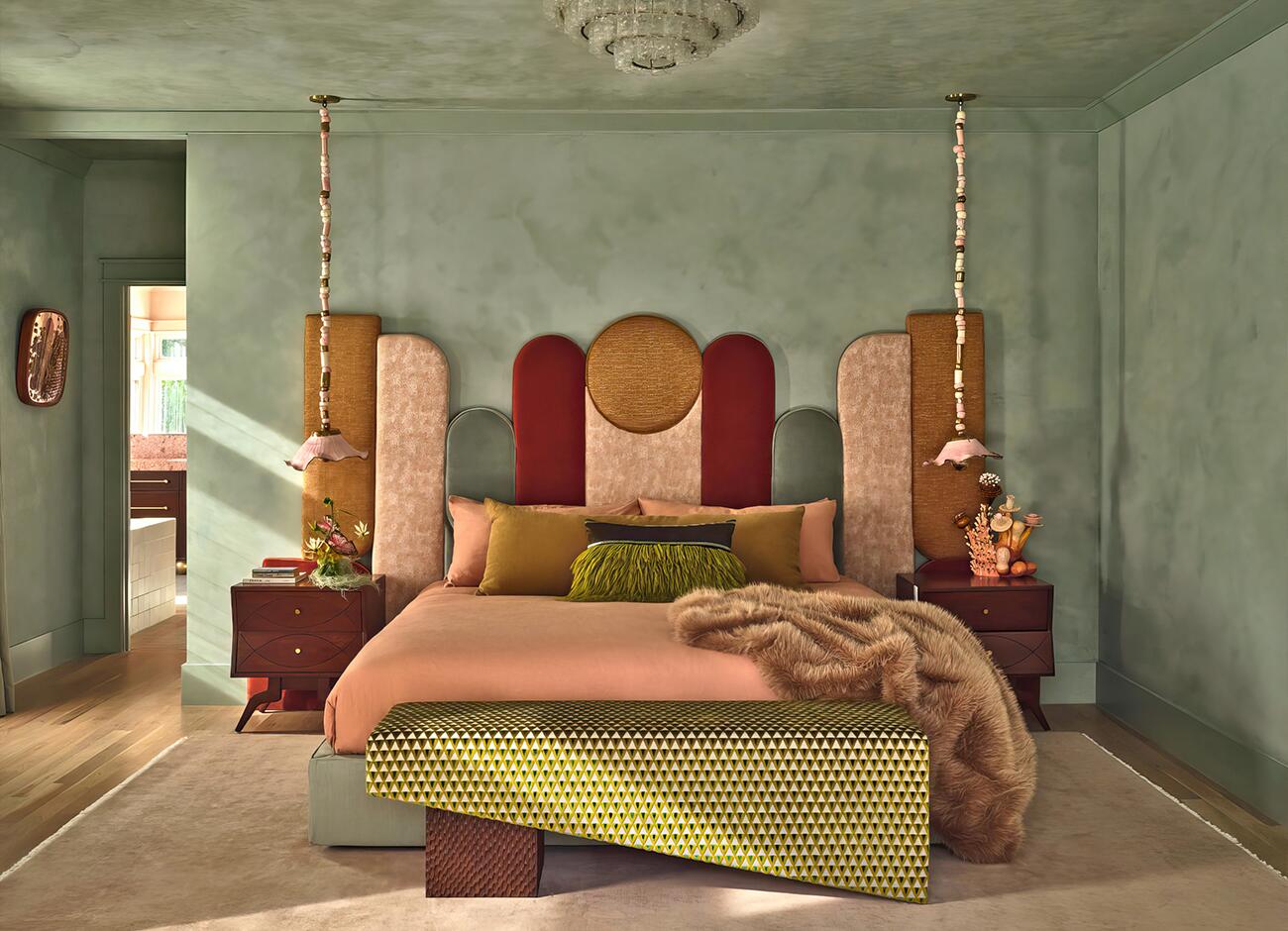 A vibrant, eclectic bedroom from a residential project by house of honey in pasadena, california. The room features the malabar ej ee bed with a tall, multi-paneled geometric headboard upholstered in a mix of kvadrat, kravet, kneedler fauchére, and zimmer + rohde fabrics in shades of ochre, burgundy, and dusty rose. The space is defined by sage green lime-wash walls and a cuff studio bench in a patterned dedar fabric at the foot of the bed. Flanking the bed are vintage nightstands illuminated by decorative sonia boyajian pendant lights with beaded cords. The styling includes a plush faux-fur throw and a textured green fringe pillow, creating a rich, layered aesthetic.