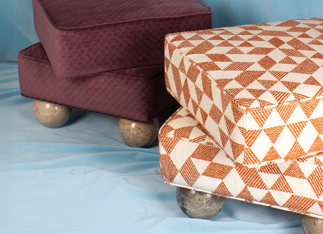 madonna-stool-12 Two Madonna Stools: one with Emperador marble sphere legs and orange Jacquard fabric; the other with Breccia marble sphere legs and purple embossed velvet fabric, set against a blue background.