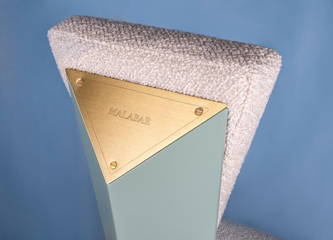 Close-up detail of the metal plate on the kju bist dining chair by malabar, featuring the malabar logo engraved into the metal, set against a blue background with the textured bouclé fabric visible.