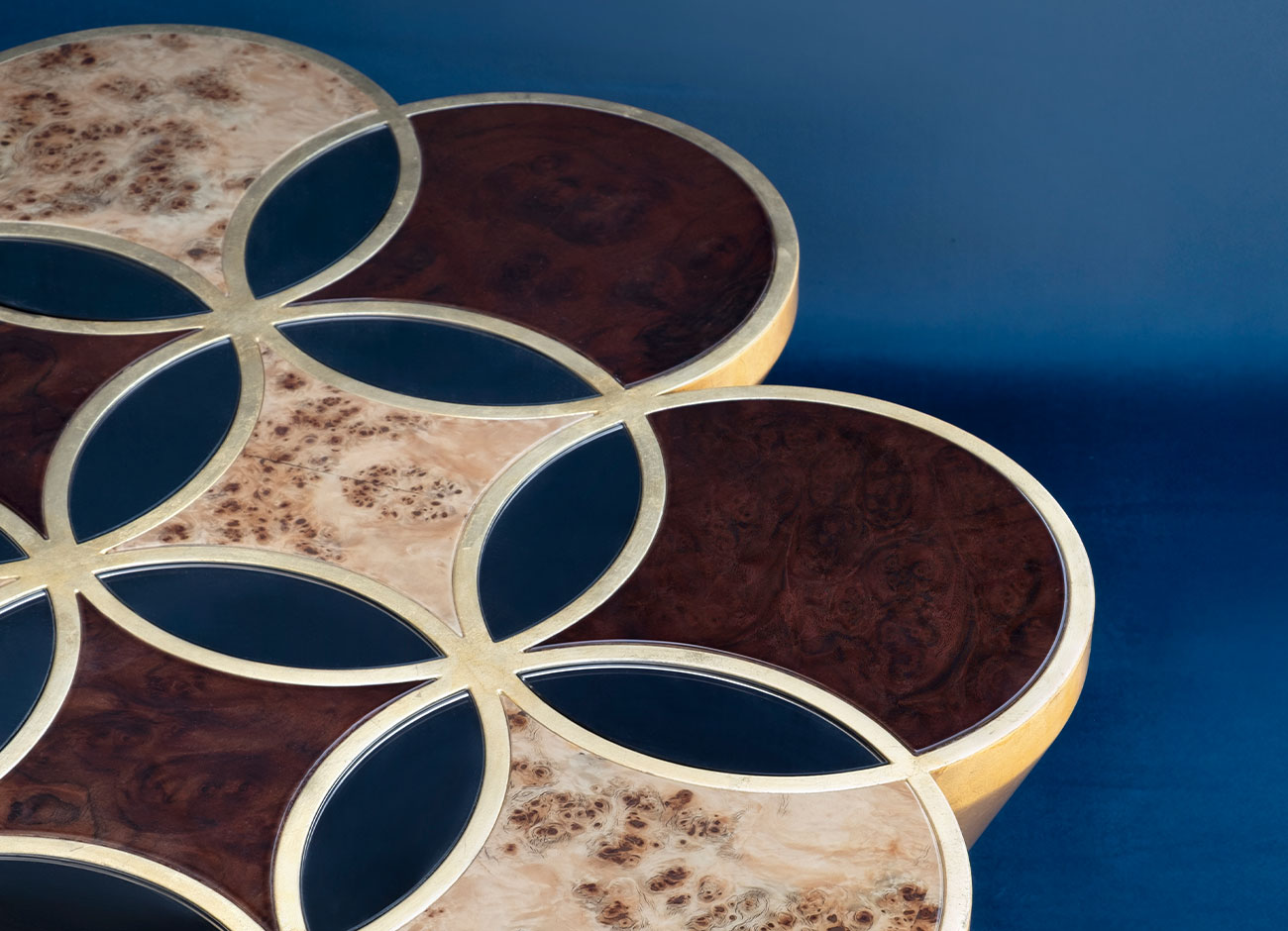 Close-up of the Ethnos center table, highlighting the textures of its gold leaf base and the walnut root and poplar root top paired with mirror, set against a rich blue background.