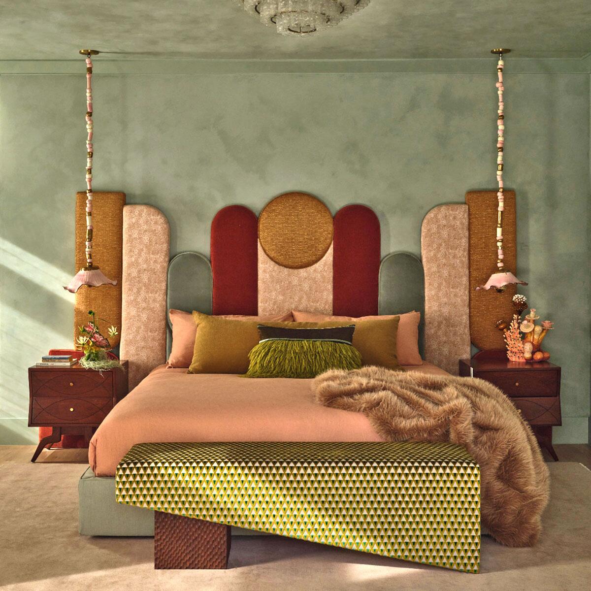 A vibrant, eclectic bedroom from a residential project by house of honey in pasadena, california. The room features the malabar ej ee bed with a tall, multi-paneled geometric headboard upholstered in a mix of kvadrat, kravet, kneedler fauchére, and zimmer + rohde fabrics in shades of ochre, burgundy, and dusty rose. The space is defined by sage green lime-wash walls and a cuff studio bench in a patterned dedar fabric at the foot of the bed. Flanking the bed are vintage nightstands illuminated by decorative sonia boyajian pendant lights with beaded cords. The styling includes a plush faux-fur throw and a textured green fringe pillow, creating a rich, layered aesthetic.