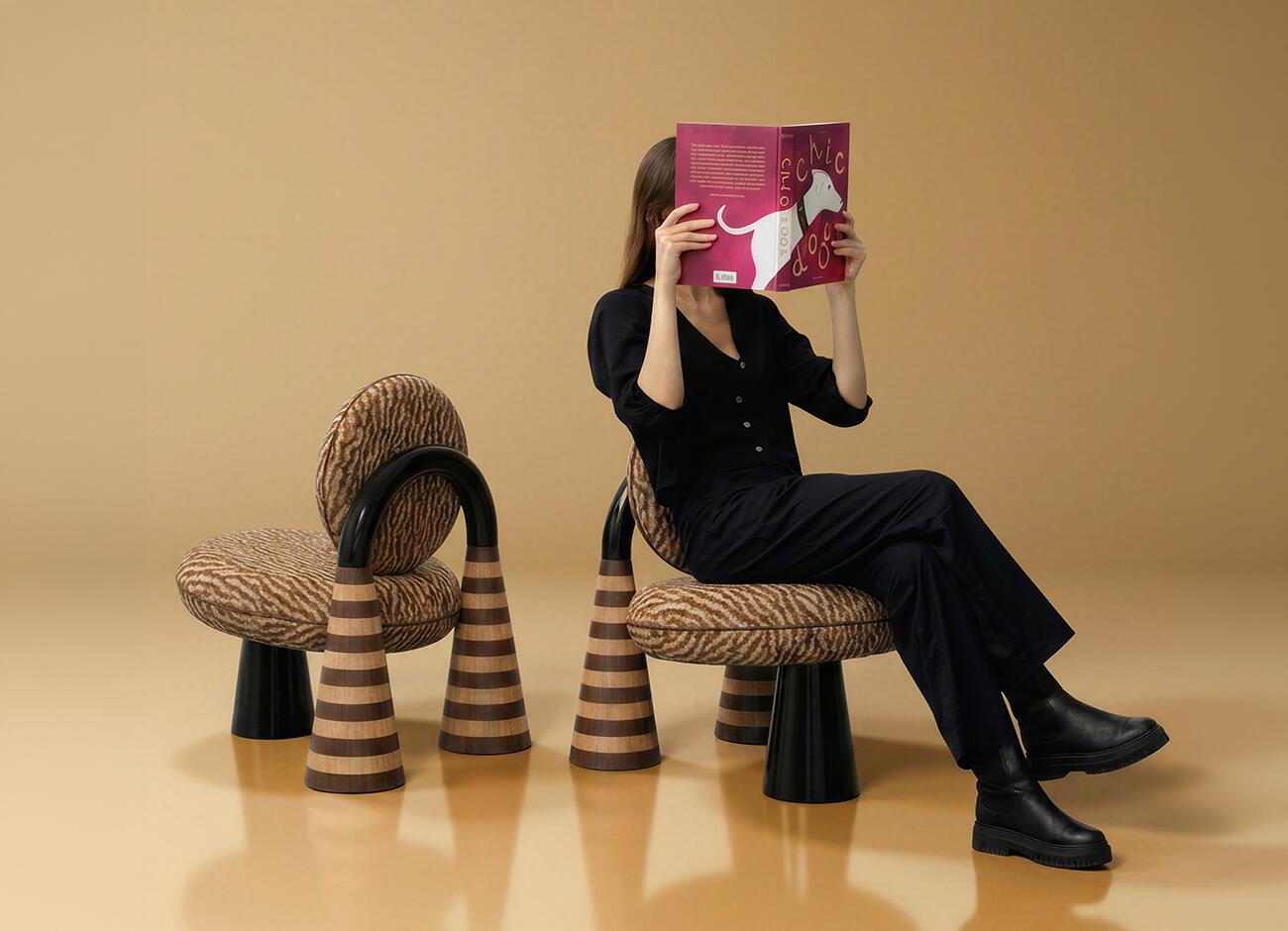 A lifestyle studio image featuring the totara armchair by malabar. A person dressed in black is seated on one armchair, holding a bright pink book in front of their face, while a second armchair is positioned next to them. The photo highlights the armchair's unique silhouette, including its patterned misia upholstery, dramatic black lacquered arch, and distinctive striped wood marquetry legs. The composition emphasizes the chair's function as both a comfortable seat and a statuesque, artistic object within a minimalist, warm-toned space.