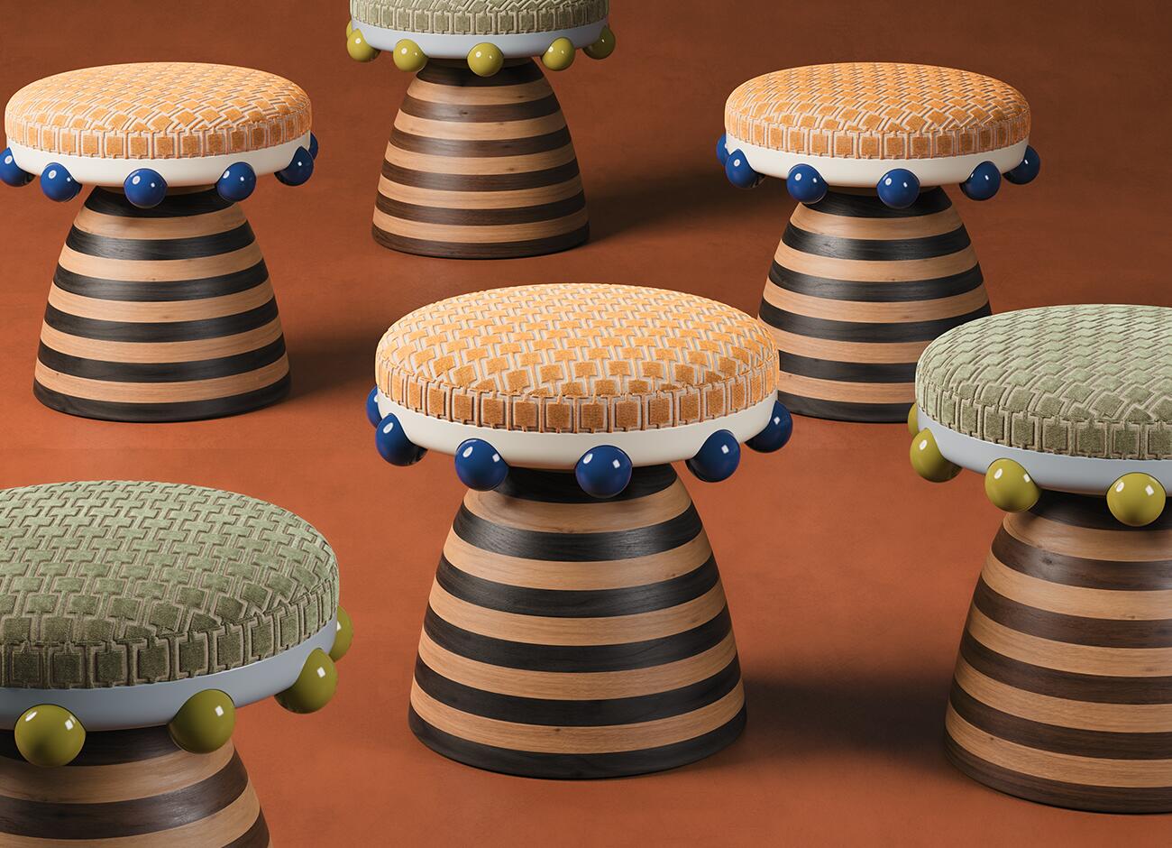 A group of krom mea stools scattered across a rust-colored floor, showcasing different colorways. Some stools feature vibrant orange patterned velvet upholstery with blue trim, while others have muted sage green velvet with lime green accents. The uniform striped wood bases create a rhythmic, graphic pattern across the frame.