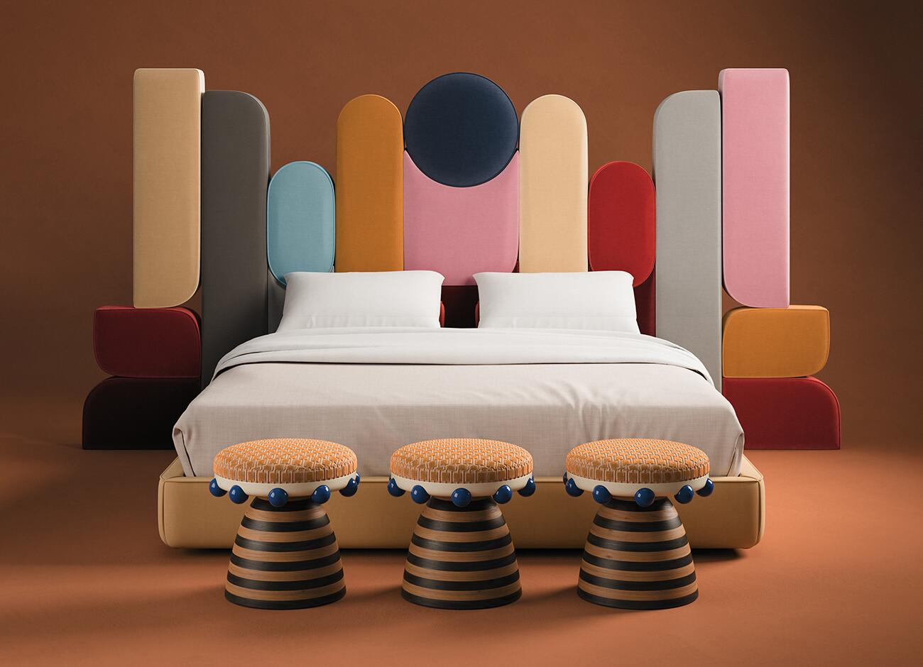 Three krom mea stools with orange patterned tops and blue spherical details standing at the foot of a bed. The bed features a massive, multi-colored headboard made of tall, rounded vertical panels in shades of pink, yellow, blue, and grey, echoing the stool's playful and expressive design language.