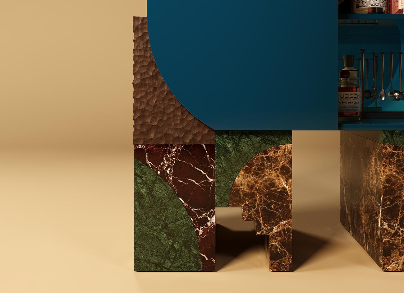 A detailed, close-up photograph focusing on the left base structure of the malabar arkos bar cabinet. The shot captures the raw texture of the hand-carved wood, contrasted by the polished, complex veins of the green (guatemala), deep red (levanto), and brown (emperador) marble segments. A glimpse of the open cabinet interior reveals stored bar tools and a bottle.