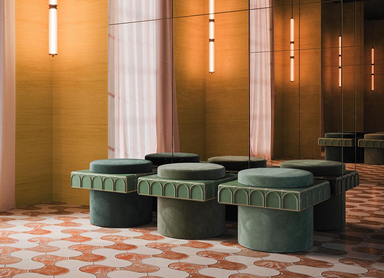 Enigma i stool A side-angle perspective photograph of three round stools arranged in a row in a richly textured interior room. Each stool features a cylindrical fabric base and a square ceramic collar with green arch-patterned tiles (barro. - emerald). The fabric seats and bases are in graduating shades of green velvet: deep forest green, olive green, and sage green, corresponding to soft jade variations. The room features a patterned floor of cream and red marble and mustard-yellow textured walls, with two floor-to-ceiling mirrors reflecting the scene and the hanging vertical light tubes. A sheer pink curtain is partially visible by a window.