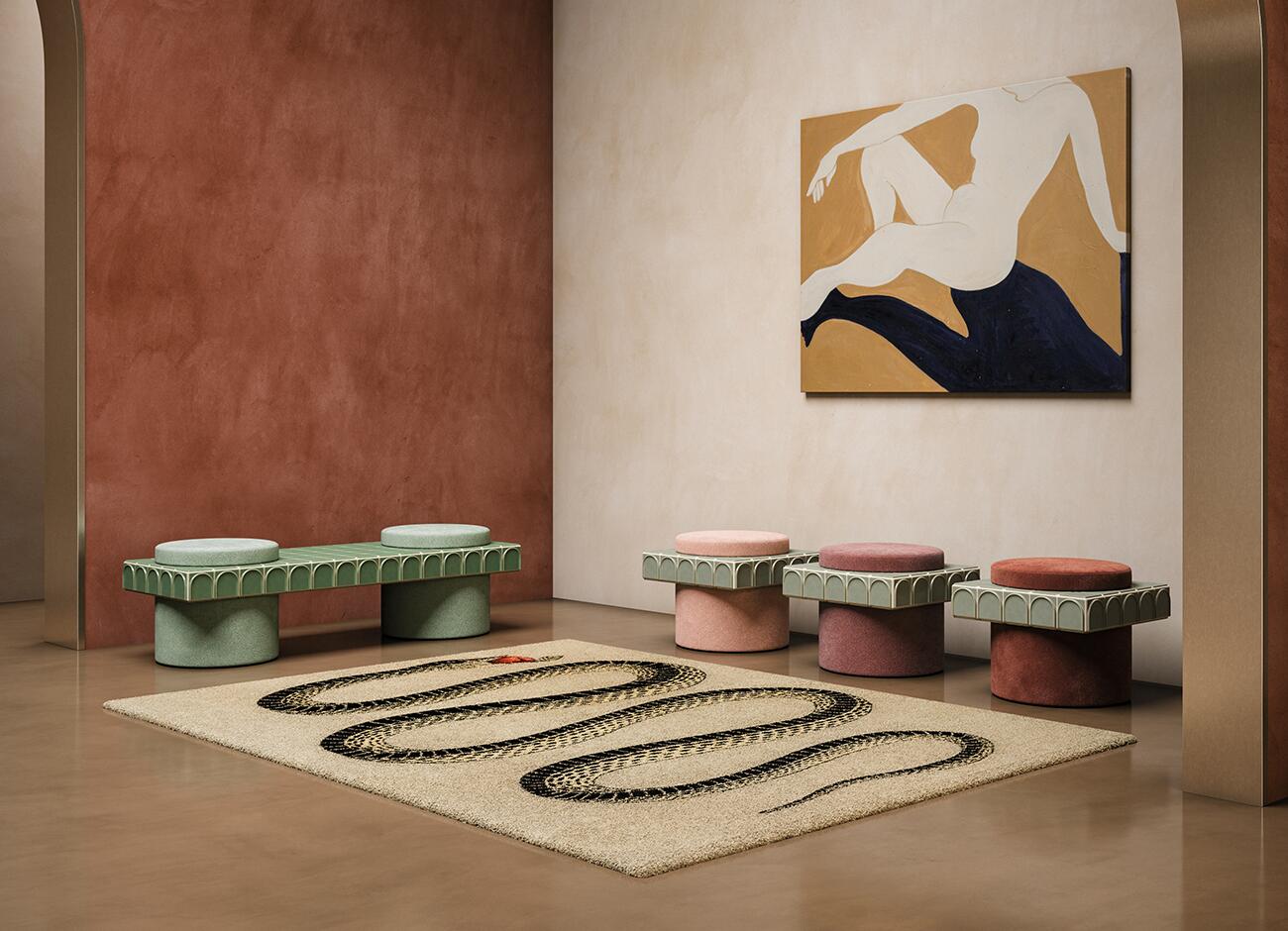 Enigma i stool A photograph of an art gallery or showroom corner with several pieces of furniture and a painting. On the left, the enigma ii stool is formed by two soft jade fabric cylindrical bases and a long connecting top surface made of light green ceramic tiles (barro. - iced) with an arched relief pattern, supporting two small round, light-green fabric seats. To the right, a row of three enigma stools stands, featuring the same green ceramic tile collar (barro. - emerald). The seats and bases are in three distinct, warm velvet shades: light pink (vintage rose), deep mauve, and rusty red (paprika). The room has a smooth, reflective brown floor and textured walls in terracotta and cream hues. Above the individual stools, a canvas painting of a stylized white nude figure against a deep blue swirl and beige background is mounted. A long, beige patterned rug with a black snake motif lies on the floor.