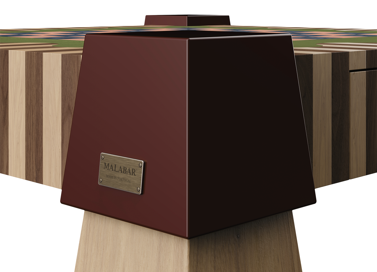 Close-up of a lacquered burgundy corner block on the Metaphorm Game Table, with a brushed aged brass plaque engraved “Malabar – Made in Portugal”, striped walnut and oak tabletop edge, and the top of a natural oak leg section below.